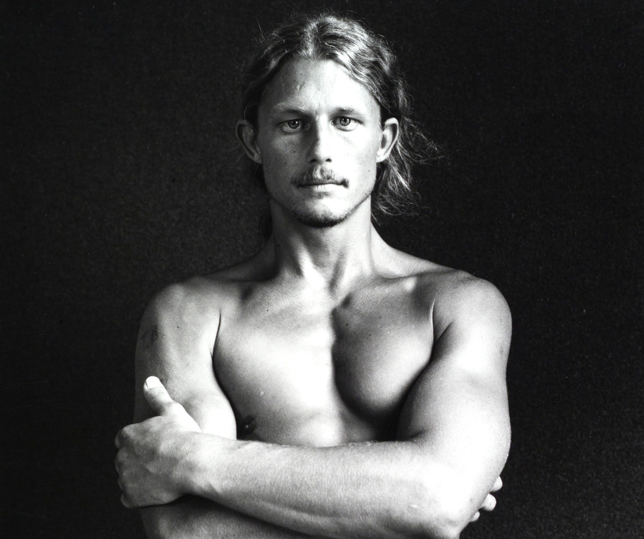 A black and white portrait of a shirtless person with long hair standing against a dark background. They have their arms crossed in front of their chest, looking directly at the camera with a serious expression.