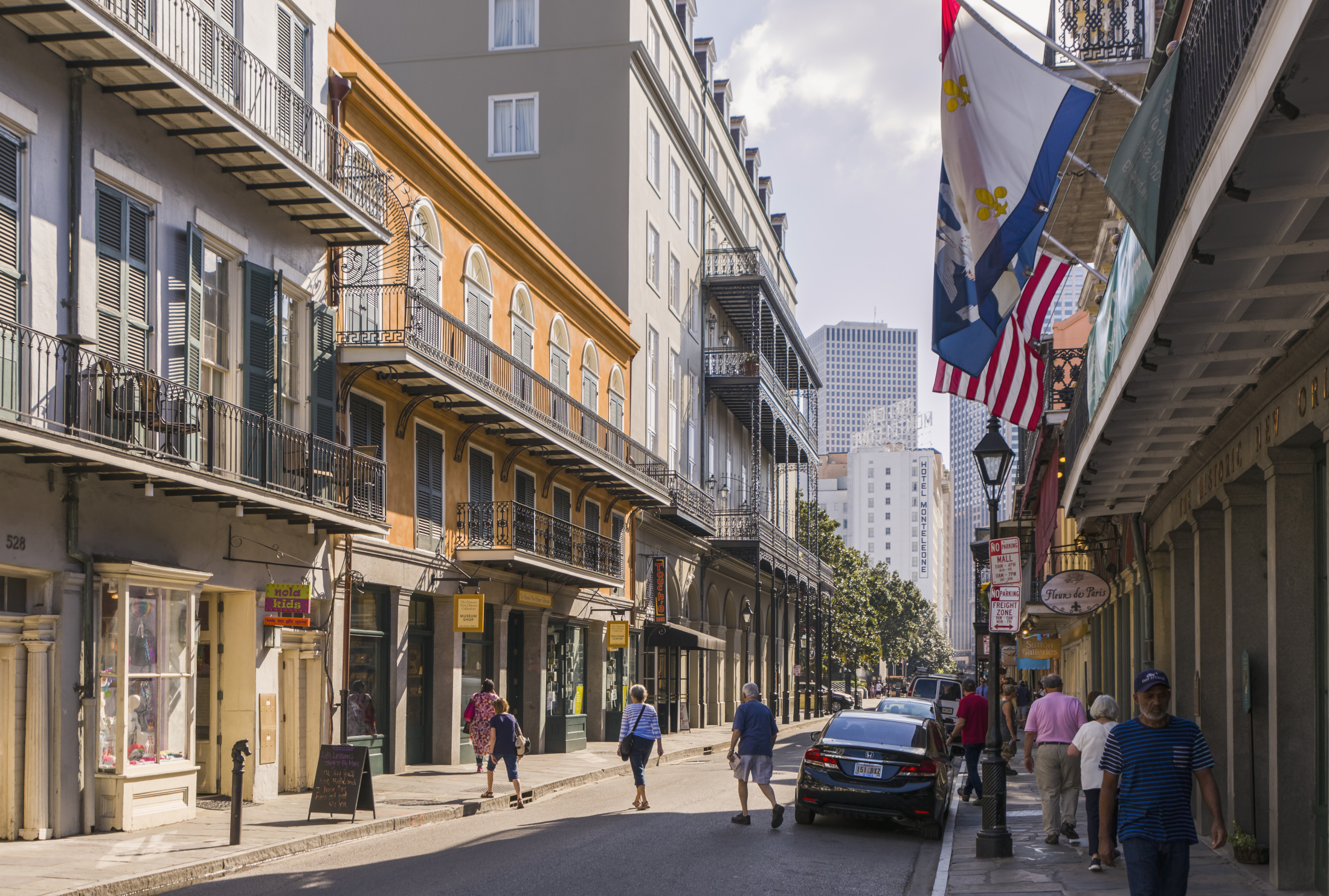 A street scene in the French Quarter of New Orleans, with people walking along a narrow road lined with buildings that have iron balconies. Theres a parked car and flags including the American and Louisiana state flag hanging above the sidewalk.