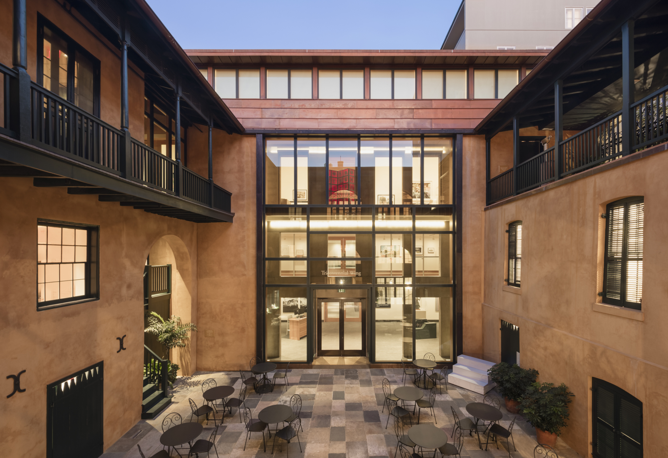 A spacious courtyard with round tables and chairs, surrounded by a historic building with large glass windows. The building has a warm, earthy exterior and balcony railings. The evening sky provides a soft, ambient light.