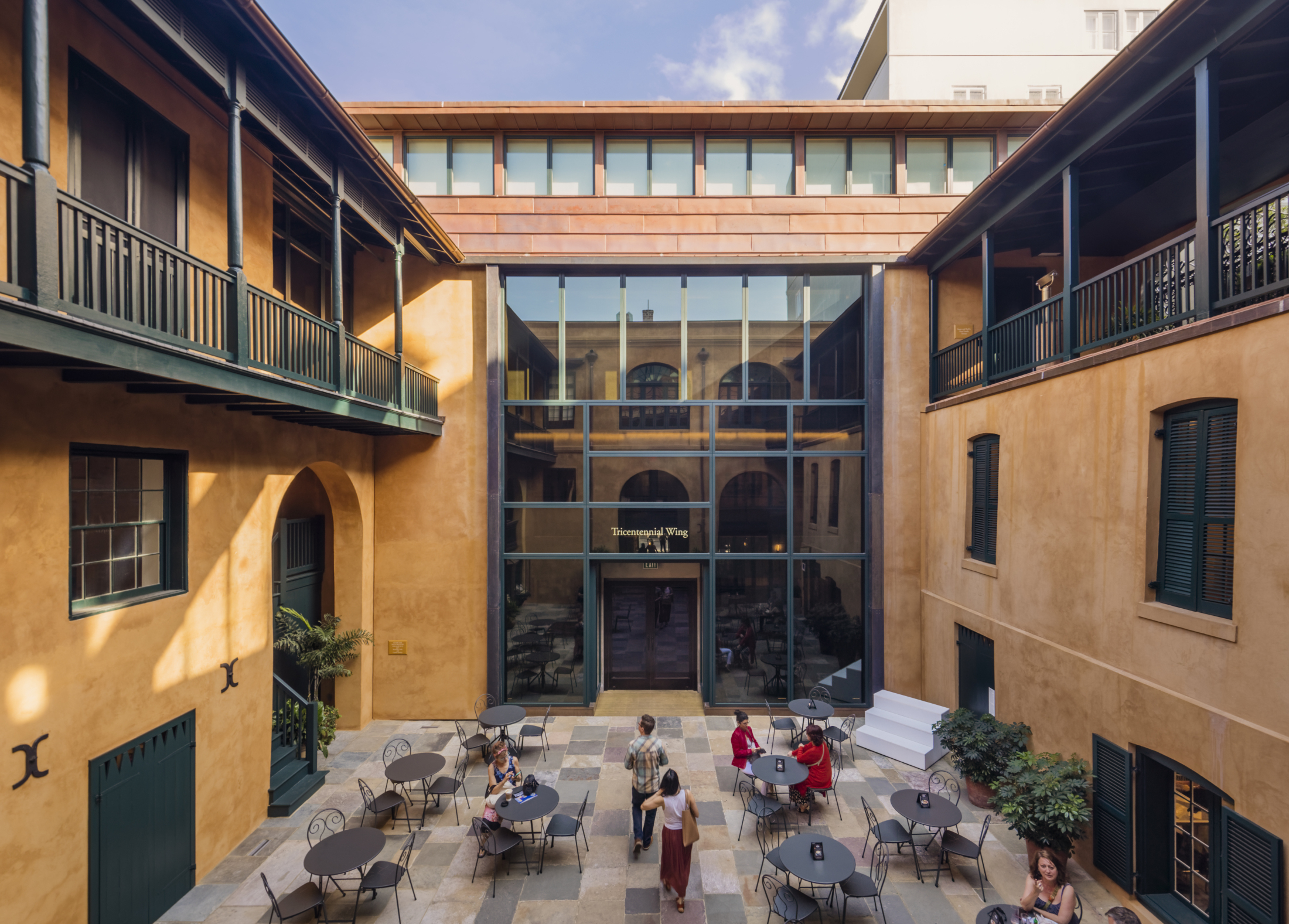 A spacious courtyard with peach-colored walls and a glass entrance. It features round tables and chairs, some occupied by people. An upper balcony stretches around the courtyard. Sunny weather and clear blue sky enhance the welcoming atmosphere.