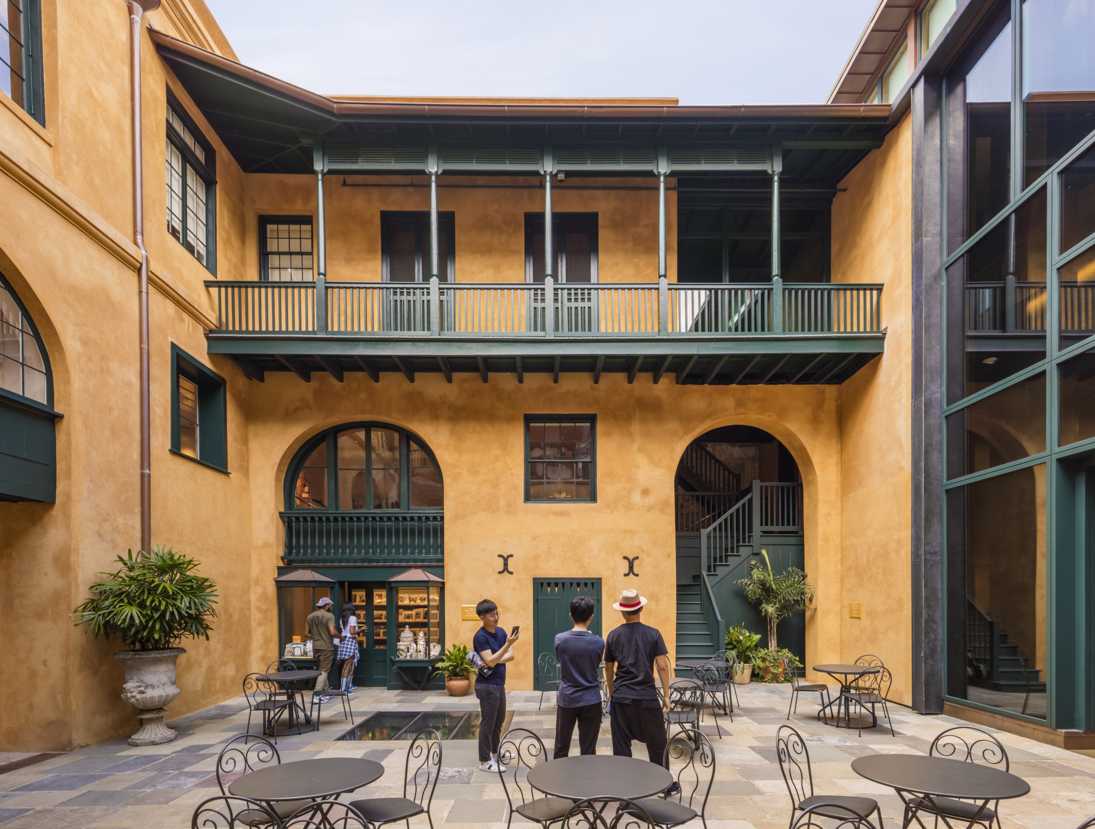 A courtyard with three people standing in the center. The building has an upper balcony with green railings and large arched windows. There are several round tables with chairs, plants, and a staircase in the background.