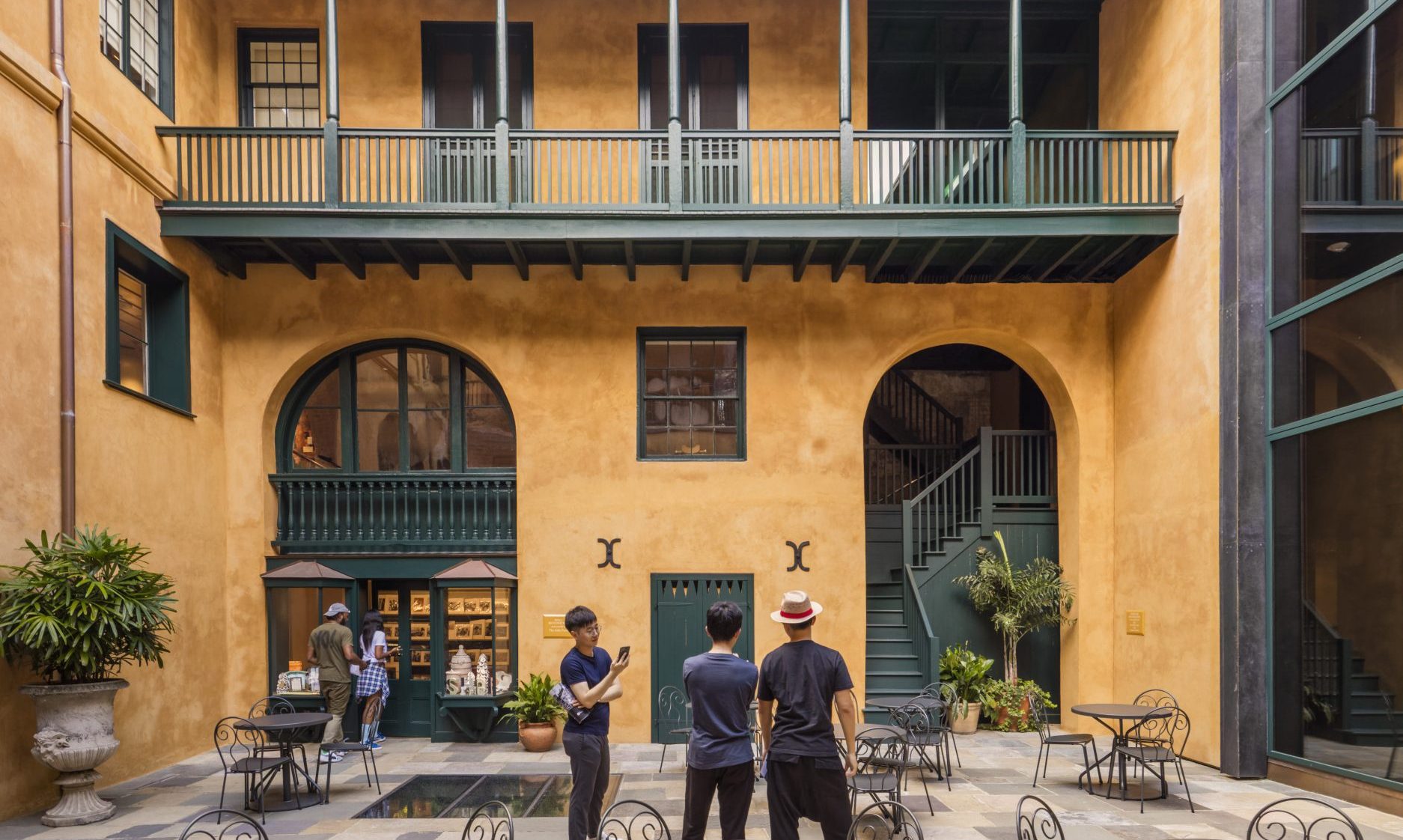 A courtyard with three people standing in the center. The building has an upper balcony with green railings and large arched windows. There are several round tables with chairs, plants, and a staircase in the background.