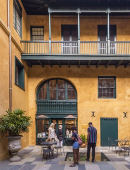 A courtyard with mustard-colored walls and dark green accents. Two people stand by a central fountain, and others browse a decorative shop with arched windows. Potted plants and wrought iron furniture complete the setting.