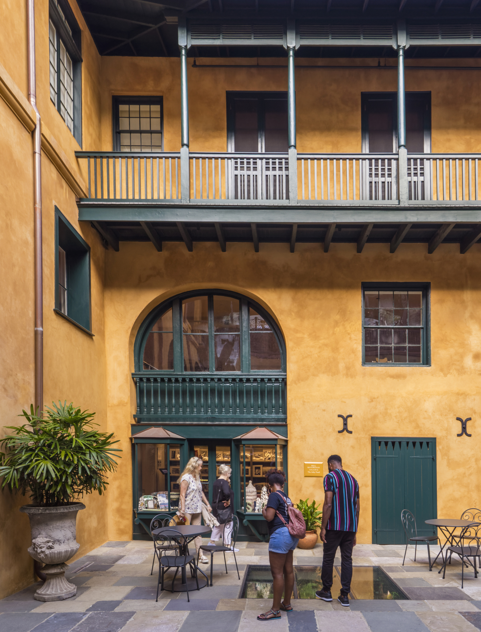 A courtyard with mustard-colored walls and dark green accents. Two people stand by a central fountain, and others browse a decorative shop with arched windows. Potted plants and wrought iron furniture complete the setting.