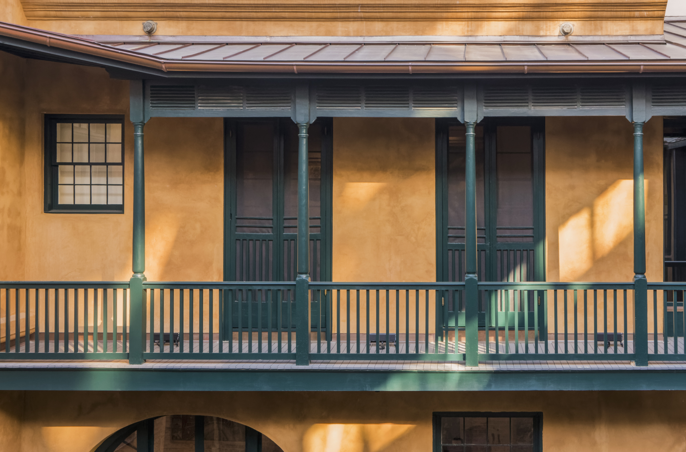 A sunlit porch with green railings and columns on the second floor of a building with tan stucco walls and a metal roof. Shadows cast across the facade, and there are windows and doors along the wall.