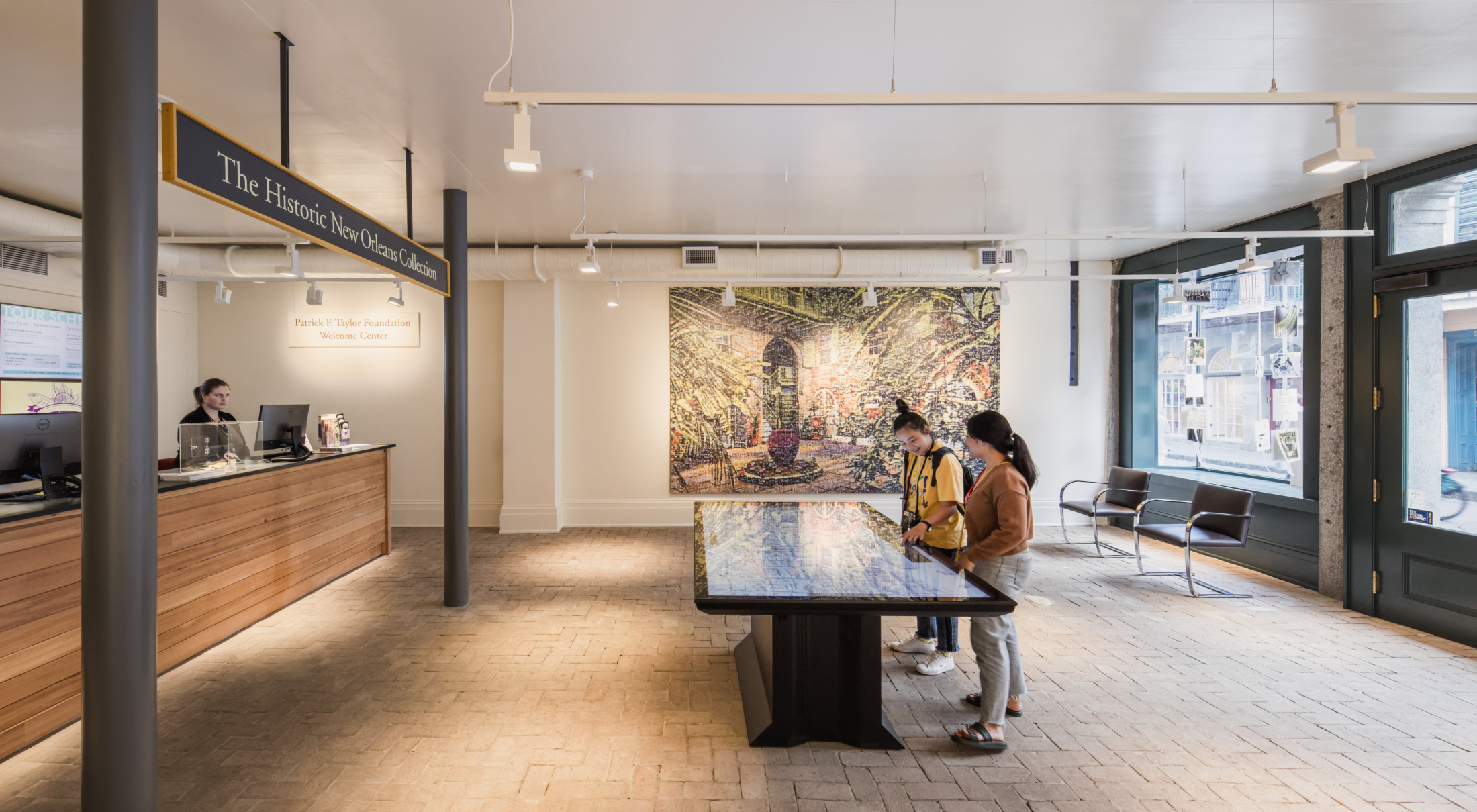 A modern lobby with two people looking at a digital display table. Behind them, a vibrant mural covers the wall. To the left, a receptionist stands behind a wooden counter. The space has a minimalist design with brick flooring and large windows.