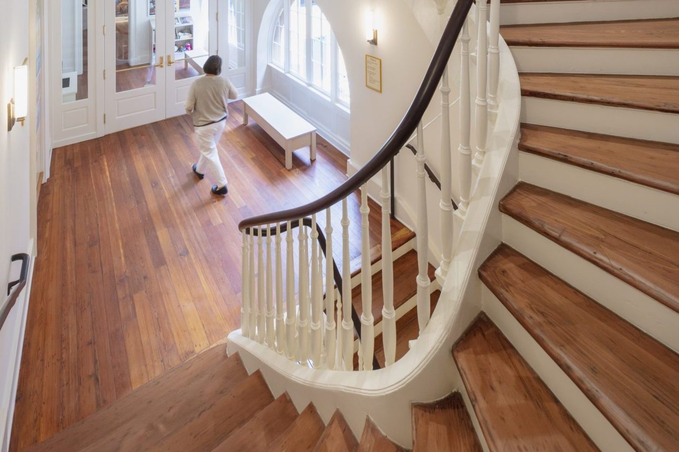 A warmly lit interior features a wooden staircase with white railing curving around a central space. A person walks below, next to large windows and an ornate gate. The walls are cream-colored, and the floor is polished wood.