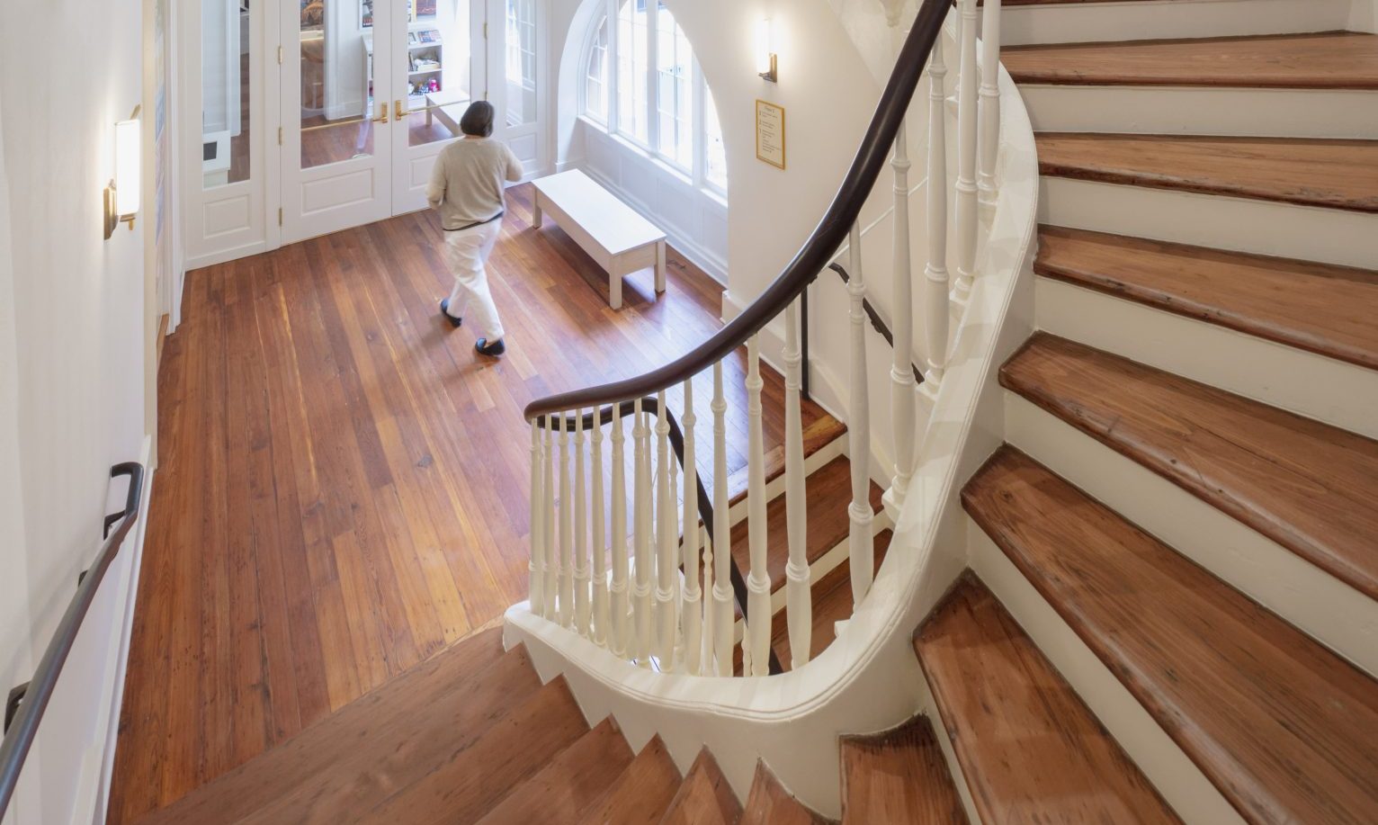 A warmly lit interior features a wooden staircase with white railing curving around a central space. A person walks below, next to large windows and an ornate gate. The walls are cream-colored, and the floor is polished wood.