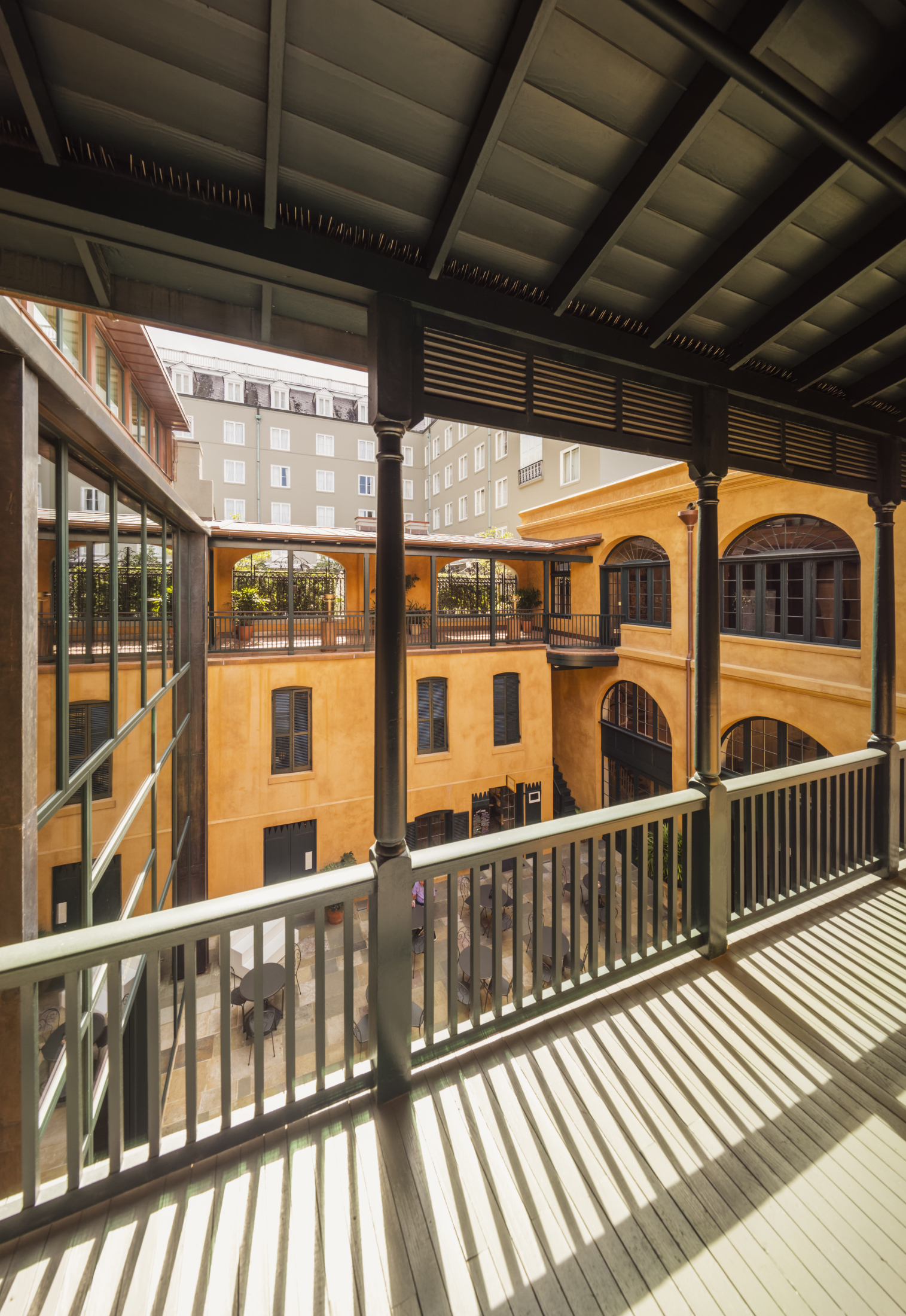 A view from a shaded balcony looking out at a courtyard surrounded by multi-story buildings with yellow walls and large windows. Shadows from the railing create patterns on the wooden floor. A few trees and tables are visible below.