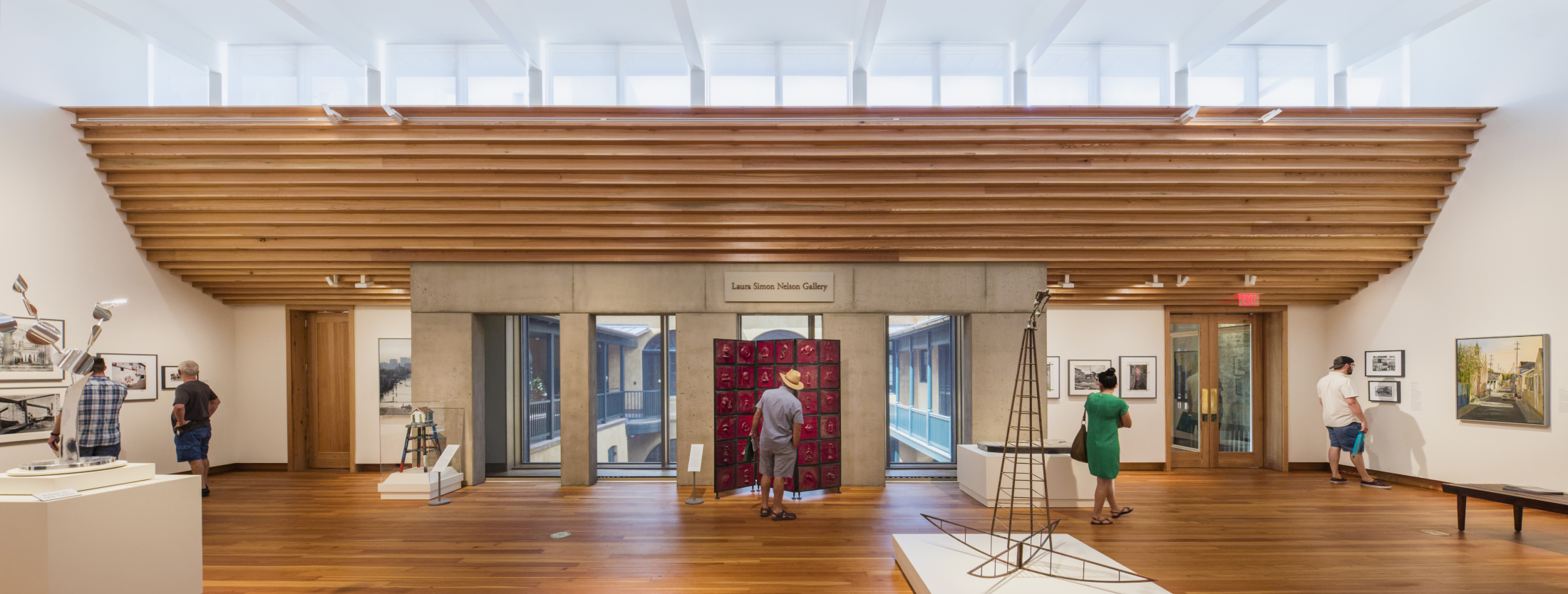 A spacious art gallery with wooden floors and ceiling beams. Several people are observing various artworks, including sculptures and framed pictures. Large windows allow natural light to flood the room, creating a bright and open atmosphere.