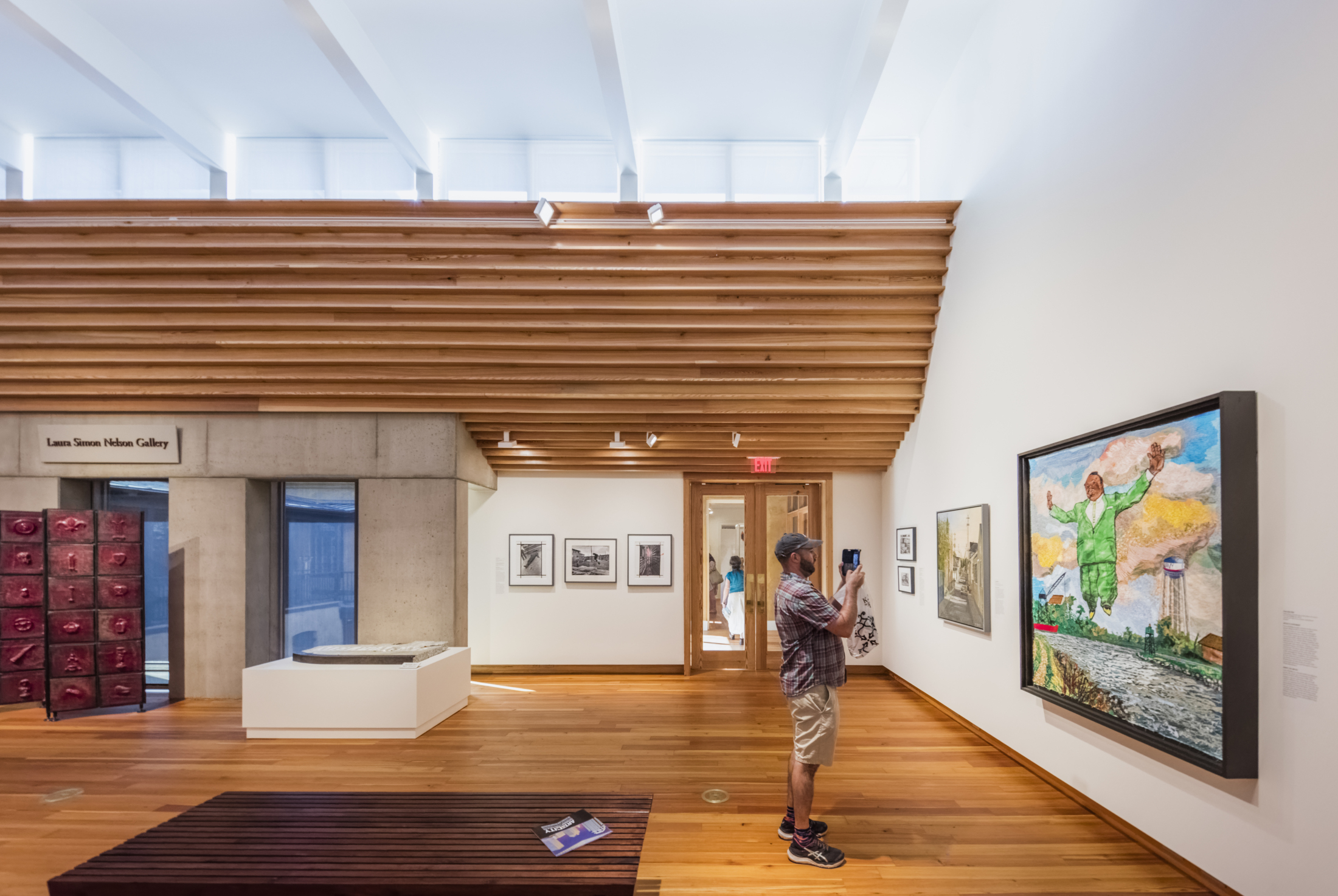 A person takes a photo of a colorful painting in a modern art gallery with wooden floors and high ceilings. The gallery walls display various artworks, and natural light streams through the windows.