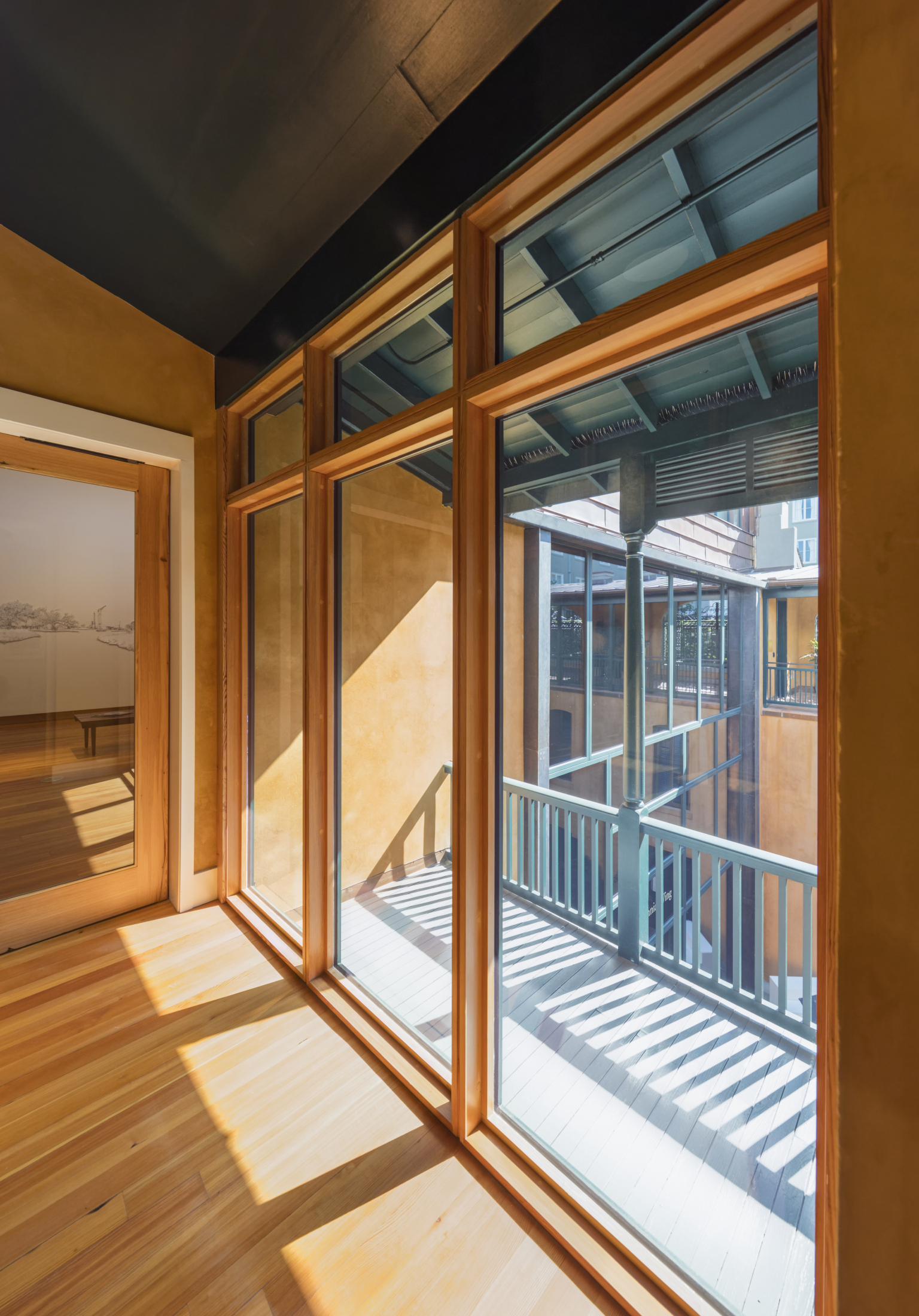 Sunlit interior with wooden floors and large floor-to-ceiling windows. The windows open to a balcony with a wooden railing. Shadows from the railing create patterns on the floor. Neutral walls add warmth to the space.