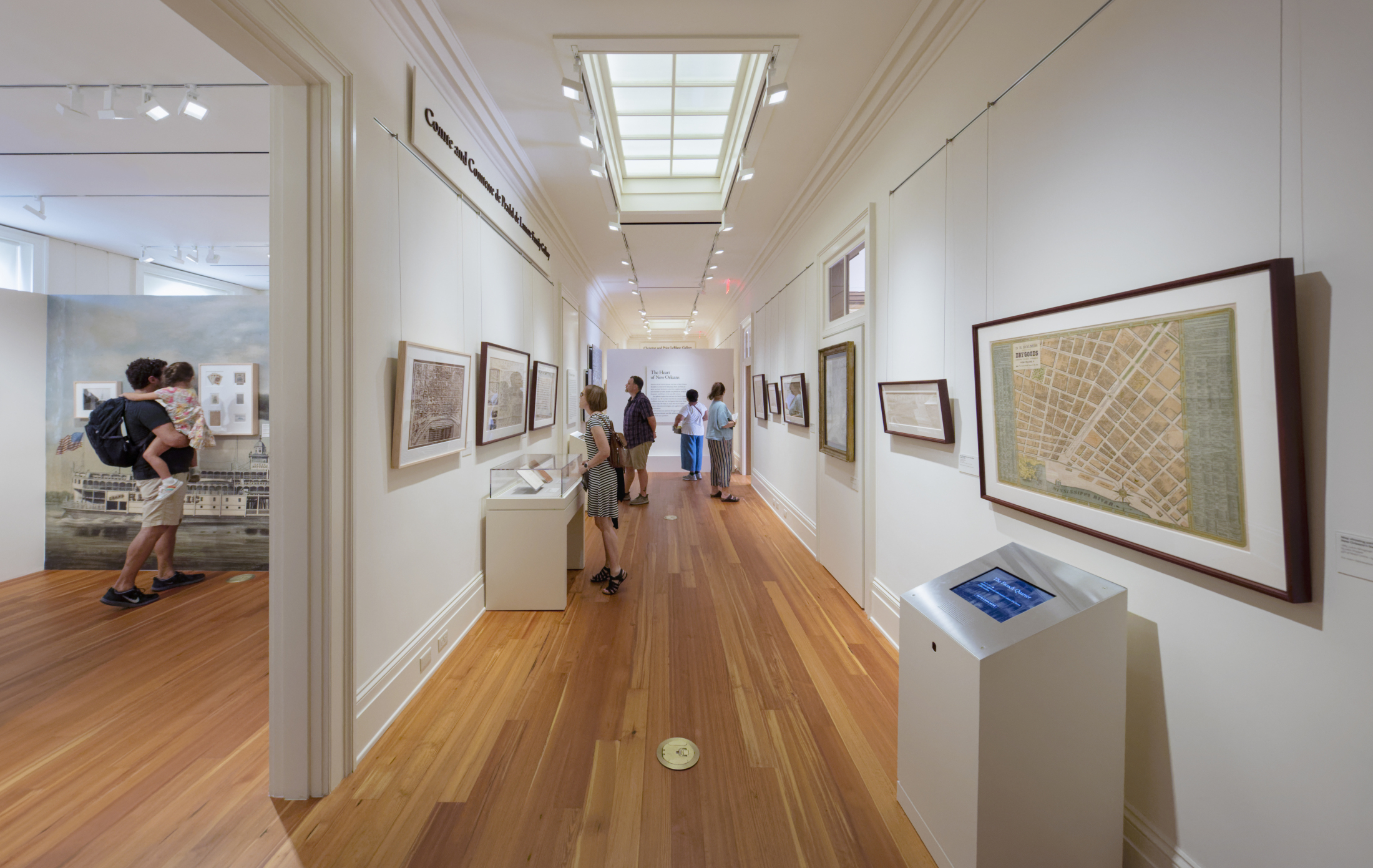 People explore a museum gallery featuring framed historical maps on white walls. The space has a wooden floor and a skylight ceiling. Some visitors examine the displays closely, while others walk through the hallway.