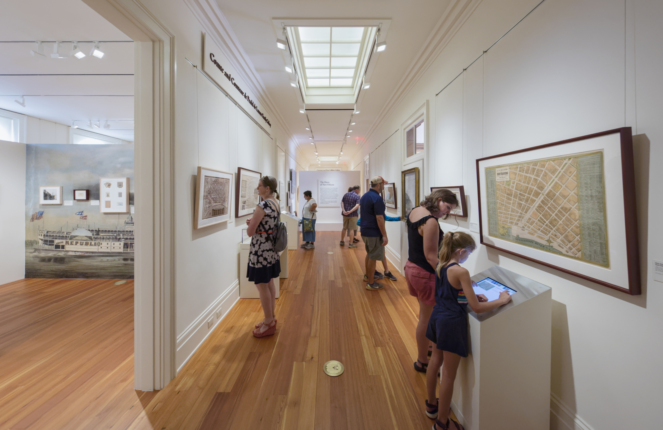 People explore an art gallery with framed maps on white walls. A child interacts with a touchscreen display. The room has a wooden floor and overhead lighting, with several visitors viewing the exhibits.