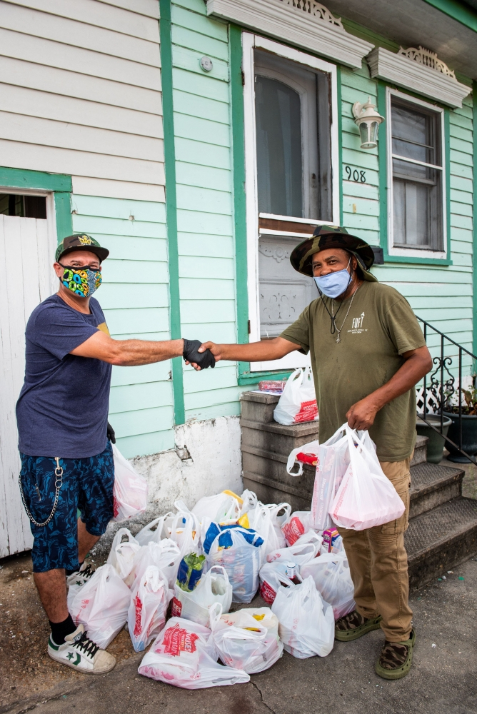 Two men wearing face masks shake hands outside a house. They are surrounded by numerous plastic bags filled with groceries on the ground. One man wears a blue shirt and shorts, the other a green shirt and beige pants.