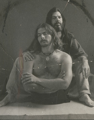 Two men with long hair pose for a vintage-style photo. The man in front is shirtless, sitting cross-legged with a serious expression, while the man behind him sits with a calm demeanor, resting his hands on the other’s shoulders.