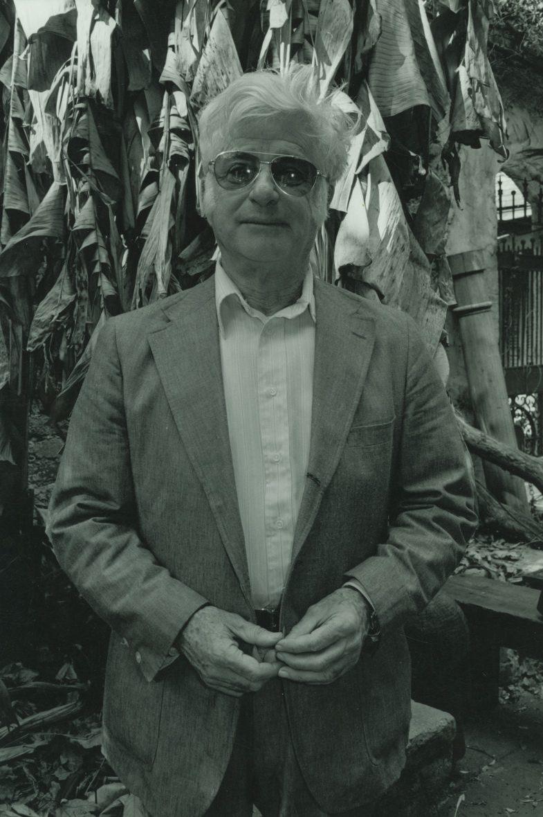 A man with white hair, wearing sunglasses and a suit, stands outdoors in front of large leafy plants.