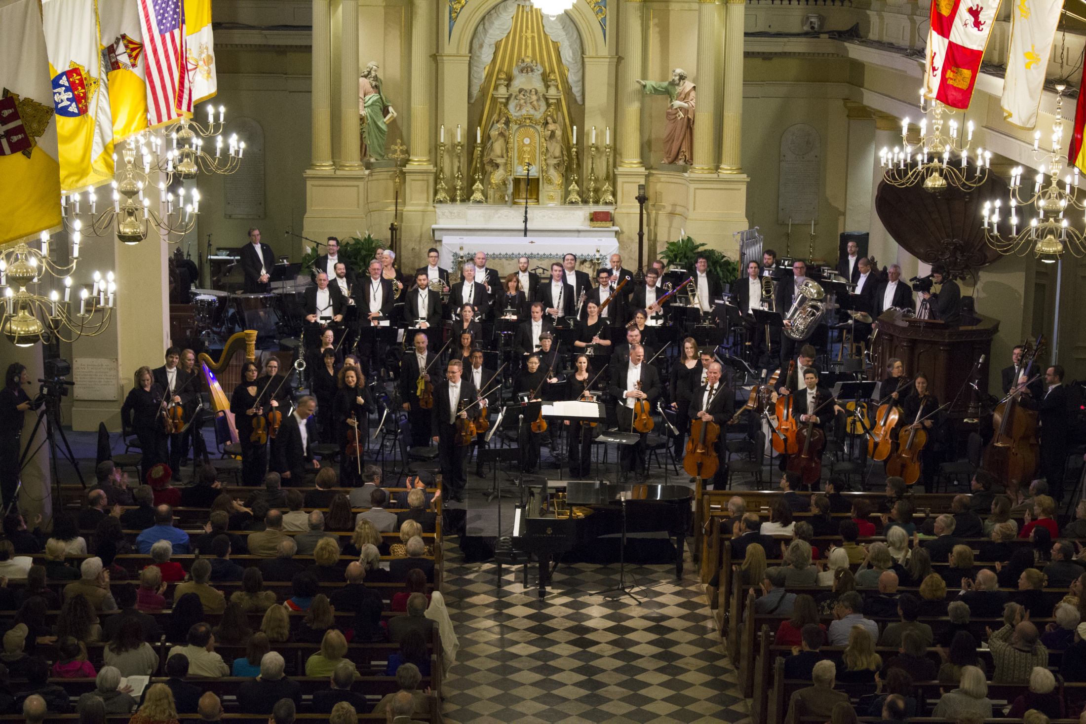 Orchestra performing in a grand church with chandeliers, colorful flags, and an ornate altar. The ensemble, in formal attire, includes various musicians and instruments. The audience, seated in rows, observes the performance.