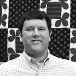 A man with short dark hair, wearing a checked shirt, smiles at the camera. The background features a black and white quilt with geometric and floral patterns. The image is in black and white.