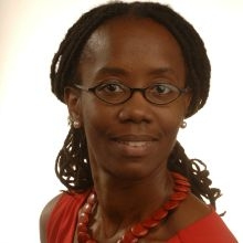 A person wearing glasses, a red top, and a red necklace is smiling slightly against a plain background. They have long, dark hair that is styled in twists.