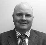 A black and white photo of a person wearing a suit and tie. The individual is bald and smiling slightly, positioned against a plain background.