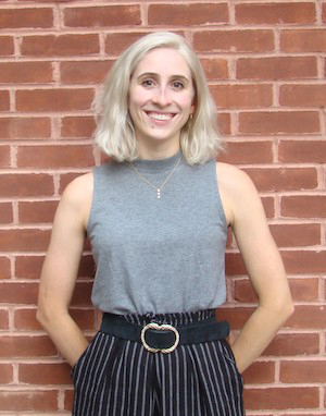 A person with shoulder-length blonde hair smiles while standing in front of a brick wall. They are wearing a sleeveless gray top and high-waisted striped pants, with hands in their pockets.