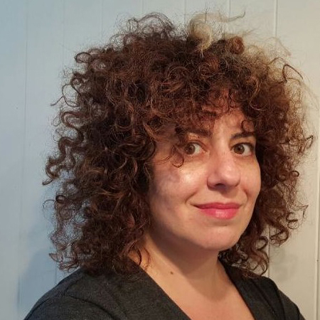 Person with curly brown hair smiling softly, wearing a dark top, standing against a light-colored background.