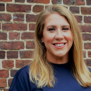 A person with long blonde hair is smiling and standing in front of a brick wall. They are wearing a dark blue top.
