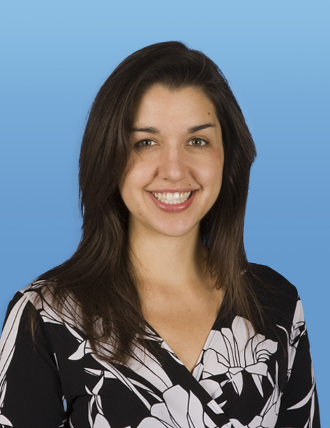 A woman with long brown hair smiles at the camera. She is wearing a black and white floral-patterned top. The background is a gradient of light to medium blue.