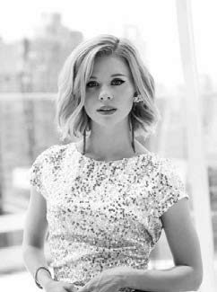 Black and white image of a woman with short, wavy hair, wearing a sequined top. She stands in front of a blurred cityscape backdrop, looking directly at the camera with a neutral expression.