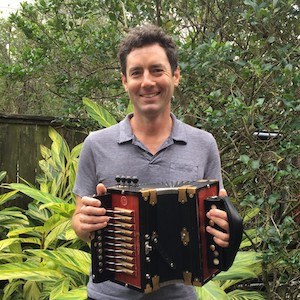 A person stands outdoors holding an accordion, surrounded by lush green plants. They are wearing a gray shirt and smiling at the camera.