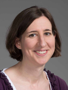 A woman with short brown hair smiles at the camera. She is wearing a dark purple cardigan over a white top against a gray background.