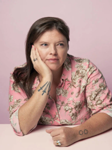 A person with long brown hair wearing a pink floral shirt rests their head on their hand. They have tattoos on their arms and a thoughtful expression. The background is light pink.