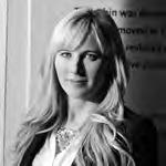 Black and white image of a person with long, light-colored hair, wearing a necklace and a dark blazer over a light shirt. They are standing indoors with a partial view of text on the wall behind them.