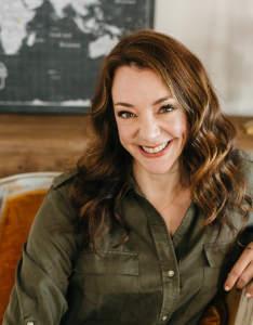 A woman with long brown hair and a green button-up shirt smiles while sitting indoors. A part of a world map is visible in the background on the wall.