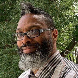 A person with glasses and a beard smiles while standing outdoors in front of green foliage. They are wearing a striped shirt.