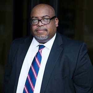 A person wearing glasses, a black suit, a white shirt, and a striped red and blue tie stands against a dark background, looking forward with a serious expression.