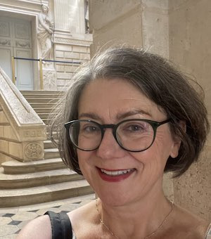 A woman with short brown hair and glasses smiles in the foreground. She stands in front of a grand staircase inside a historic building with ornate stonework and large doors.