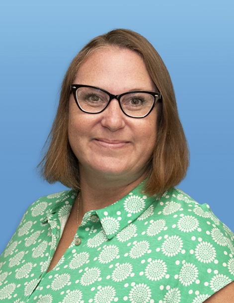 A woman with shoulder-length brown hair and glasses smiles at the camera. She is wearing a green blouse with a white floral pattern. The background is a gradient of light to medium blue.
