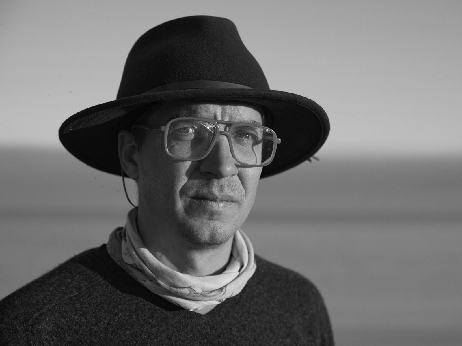 A black and white portrait of a person wearing large glasses, a wide-brimmed hat, and a scarf. The background is blurred, suggesting an outdoor setting, possibly a beach or open landscape. The person gazes calmly at the camera.