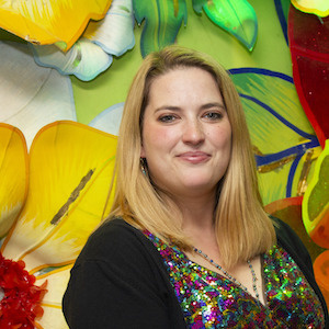 A woman with blonde hair is smiling in front of a colorful, floral mural. She is wearing a multicolored, sparkly top and a black cardigan.