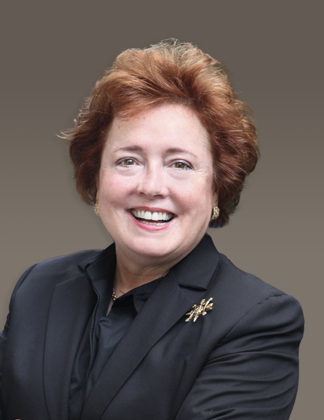 A woman with short, curly, auburn hair is smiling at the camera. She is wearing a black blazer with a gold brooch on the lapel and a black shirt. The background is a neutral gray-brown gradient.