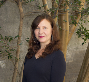 A woman with long brown hair is standing in front of a tree with branches visible in the background. She is wearing a dark long-sleeve shirt and hoop earrings, smiling gently at the camera.
