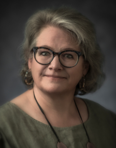 A woman with glasses and shoulder-length gray hair, wearing a green top and earrings, poses against a dark background.