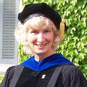 A person with curly blond hair, wearing a graduation cap with a gold tassel and a gown with a blue velvet hood, smiles in front of a leafy background.