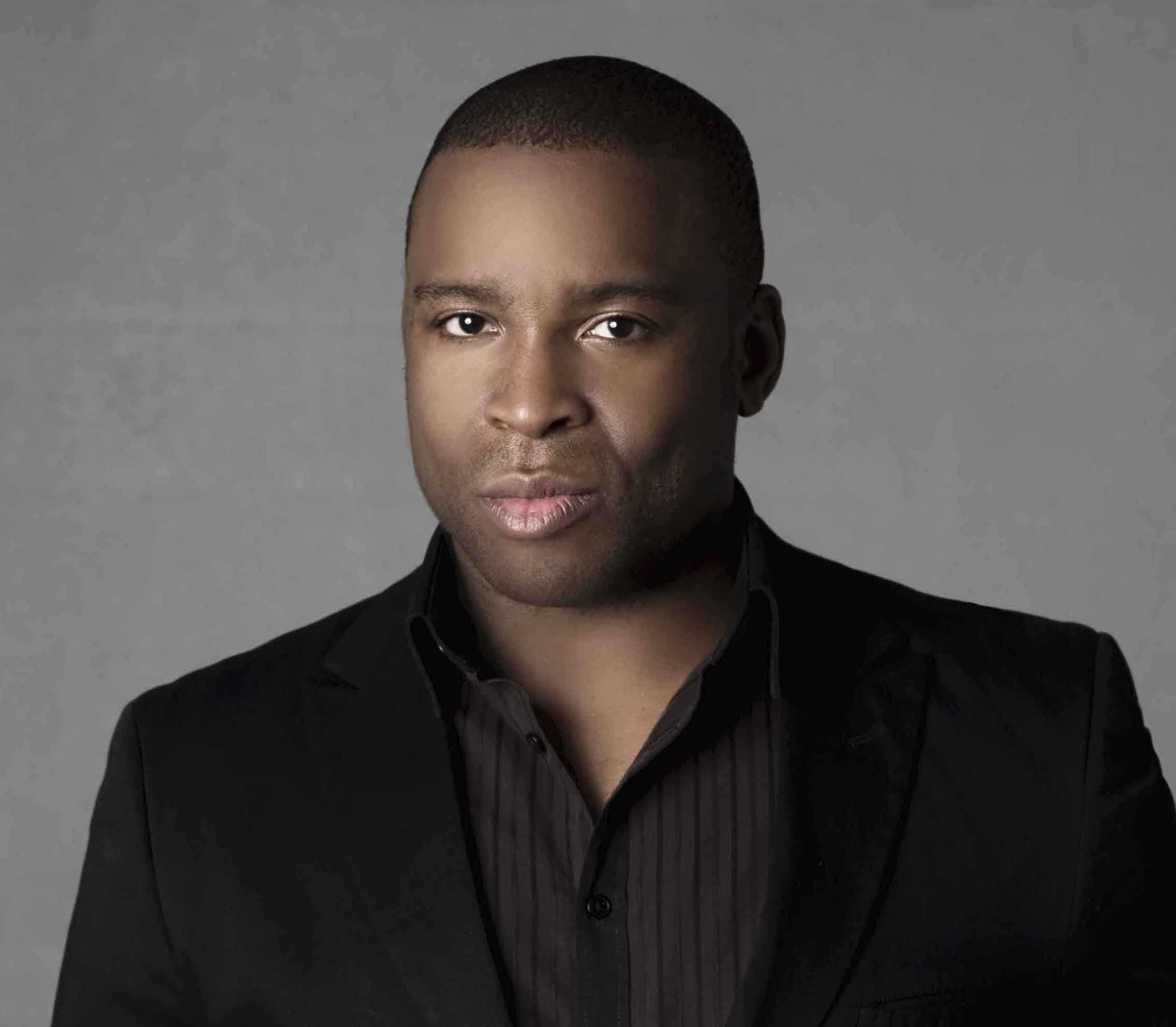 A person with a neutral expression wearing a black blazer and shirt poses against a gray background.