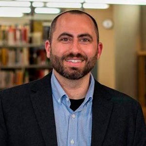 A person with a beard wearing a blue shirt and dark blazer is smiling. They are standing indoors with bookshelves visible in the background.