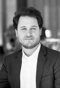 Black and white photo of a person wearing a suit jacket over a white shirt, looking towards the camera with a neutral expression. The background is blurred, suggesting an indoor setting.