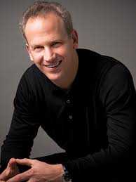 A person with short hair, wearing a black long-sleeve shirt, is smiling while leaning slightly forward. The background is a simple gray.