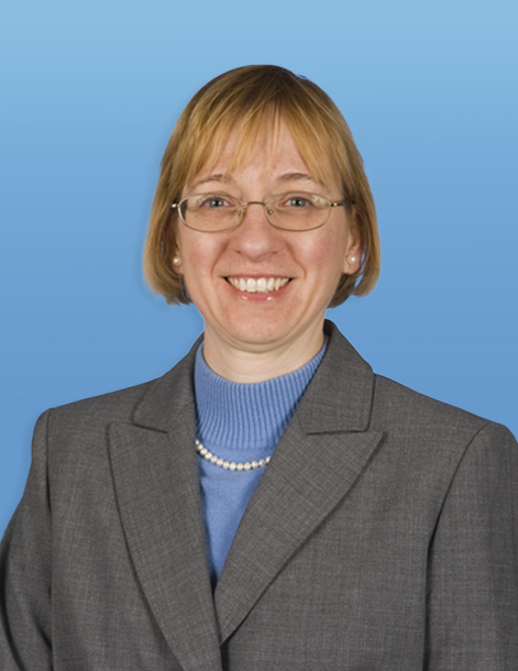 A smiling person wearing glasses, a pearl necklace, a blue turtleneck, and a gray blazer against a solid blue background.